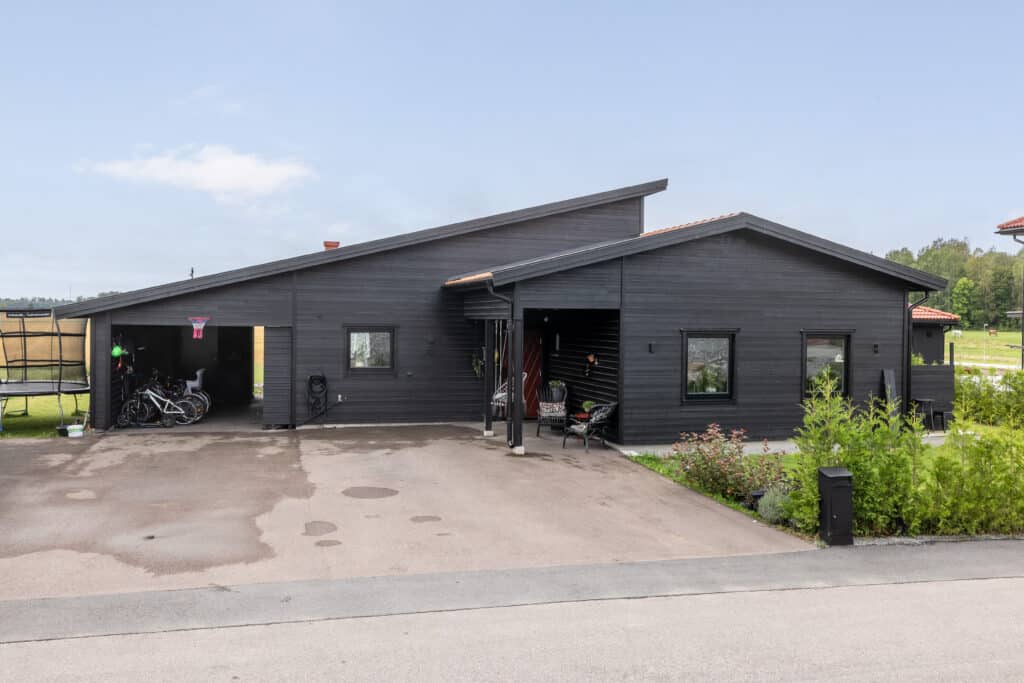Exteriörbild av ett mörkmålat trähus med asfalterad garageuppfart, cyklar i en öppen carport och en uteplats vid entrén.
