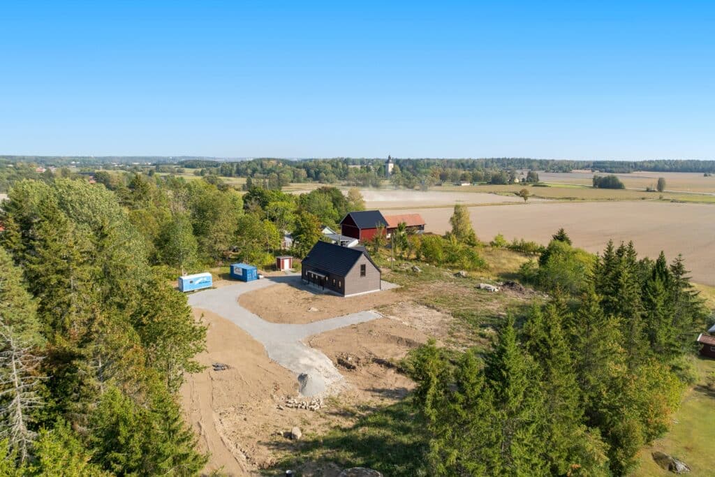 Flygfoto över ett modernt hus under uppbyggnad på en tomtyta i ett lantligt landskap med ängar och skog, vilket illustrerar tomtplanering och husplacering.